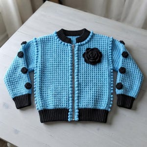 May include: A light blue crochet bomber jacket featuring a textured waffle stitch pattern, a central silver zipper, and black ribbed trim at the collar, cuffs, and hem. The jacket is decorated with small black crochet flowers and blue bobbles.