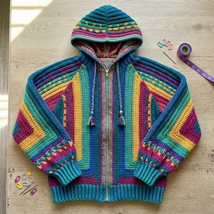 May include: A zip-up hooded jacket featuring a multi-colored crochet pattern in shades of teal, magenta, yellow, and blue. The interior lining displays a paisley print. Two braided drawstrings with wooden beads hang from the hood.