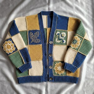Könnte beinhalten: Eine langärmelige Häkelstrickjacke mit einem Patchwork-Design aus cremefarbenen, senfgelben, waldgrünen und marineblauen Quadraten. Das Kleidungsstück hat vier dunkle Knöpfe, einen gerippten marineblauen Saum sowie gestickte Blumen- und Blattmotive.