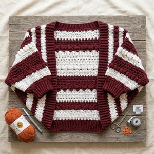 May include: A crochet cardigan featuring horizontal stripes in deep burgundy and white, with textured bobble stitch patterns. The garment is displayed with a ball of orange yarn, a crochet hook, scissors, stitch markers, and a 36-inch measuring tape.