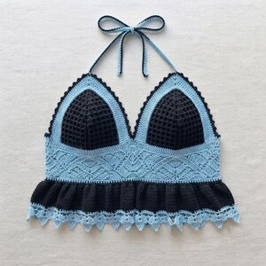 Puede incluir: Un top halter de ganchillo con copas triangulares negras en punto de gofre, bordeadas con hilo azul claro. La sección media tiene un intrincado diseño de encaje azul claro, terminado con un dobladillo negro con volantes y un borde festoneado.