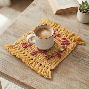 May include: A square yellow crocheted coaster with fringe edges, featuring a dark pink floral and vine embroidery pattern. A speckled ceramic mug filled with coffee and a small gold-toned metal spoon are placed on top of the textured fabric mat.