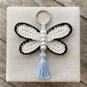 May include: A dragonfly-shaped keychain featuring cream-colored crocheted wings with black borders. The central body is made of light blue yarn with five white spherical beads arranged vertically, topped with a silver metal ring and a light blue tassel.