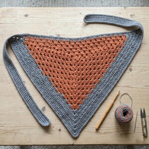 Puede incluir: Un chal triangular de ganchillo con una sección central de hilo naranja rodeada por un borde gris. El chal tiene lazos largos en cada extremo. Junto al chal hay un ovillo de hilo naranja y gris, un ganchillo de madera, una aguja y unas tijeras.