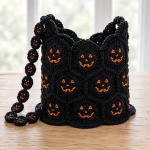 Pode incluir: Uma bolsa de crochê preta em formato de balde, construída com motivos hexagonais, cada um apresentando um rosto de abóbora bordado em laranja. A bolsa inclui uma alça combinando feita de pequenas peças circulares de crochê preto com detalhes laranja.