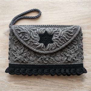 May include: A gray crochet clutch bag featuring a textured floral pattern and a black six-pointed star motif on the flap. The bag includes a matching gray wrist strap and a decorative black scalloped border along the bottom edge of the purse.