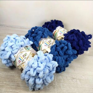 May include: Four skeins of Alize Puffy yarn in varying shades of blue, including light blue and dark navy. Each skein features a pre-looped, soft, textured design with a paper label wrapped around the centre displaying the Alize Puffy brand name.