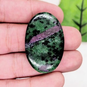 Può includere: Una gemma di zoisite di forma ovale con una superficie lucida caratterizzata da un motivo maculato di verde bosco profondo, inclusioni nere sparse e una prominente striscia allungata di rubino color magenta vivido al centro.