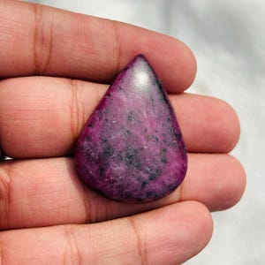 May include: A teardrop-shaped ruby zoisite gemstone featuring a vibrant magenta and deep pink base color with scattered dark green and black mineral inclusions throughout the polished, smooth surface of the stone.