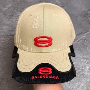 May include: A beige baseball cap with distressed detailing, featuring a red embroidered number eight logo on the front panel. The brim is layered with a black accent piece that displays a red number eight logo and the text BALENCIAGA in red.