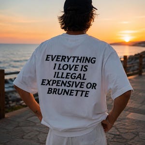 May include: A white short-sleeved t-shirt featuring black text on the back that reads: EVERYTHING I LOVE IS ILLEGAL EXPENSIVE OR BRUNETTE. The shirt is worn with white shorts and a black baseball cap, viewed from the back against a sunset background.