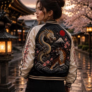 Peut inclure: Un blouson bomber en satin noir et crème avec une broderie complexe au dos. Le motif représente un dragon, un tigre, une montagne, un soleil rouge et des branches de cerisier. Les manches crème présentent des broderies florales et des poignets rayés.