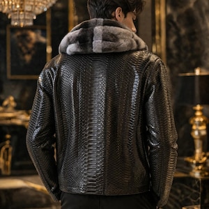 Peut inclure: Une veste noire avec un motif texturé en peau de serpent et une capuche doublée de fourrure grise et blanche. La veste présente des manches longues et un ourlet droit, portée sur un pantalon sombre. Le matériau a une finition brillante.