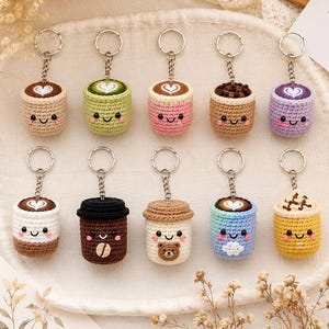 May include: Ten crocheted keychains shaped like small cups with smiling faces. Each cup features unique details like latte art, coffee beans, a bear face, or whipped cream toppings in various colors including pink, green, purple, blue, and brown.