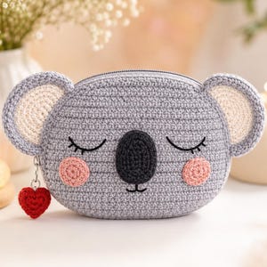 Op de afbeelding: Een grijs gehaakt koala-tasje met een donkergrijze neus, gesloten ogen met wimpers en roze ronde wangen. Het tasje heeft afgeronde oren met crèmekleurige centra en een kleine rode hartvormige bedel bevestigd aan de ritssluiting.