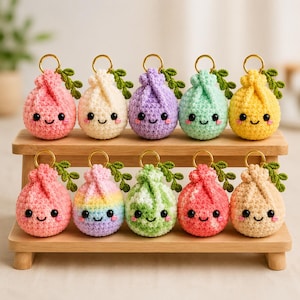 May include: Ten round, crocheted plush keychains arranged on a wooden stand. Each keychain features a smiling face with black eyes, pink cheeks, and a small green crocheted leaf attached to a gold metal ring. The colors include pink, white, purple, mint, yellow, rainbow, green, and peach.