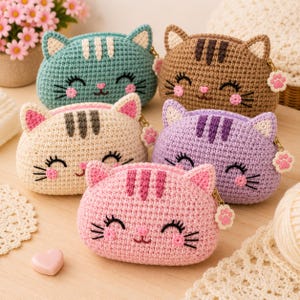 May include: Five small, crocheted cat-shaped pouches in teal, brown, cream, purple, and pink. Each pouch features embroidered facial details, including closed eyes, whiskers, pink cheeks, and a small nose, with a cat paw charm on the zipper pull.