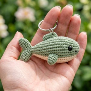 May include: A small, handmade crochet fish keychain featuring a sage green upper body and a cream-colored underbelly. The fish has a textured yarn surface, a single dorsal fin, a tail fin, a side fin, and two small black plastic bead eyes.