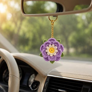 Peut inclure: Une décoration florale en crochet suspendue à une chaîne en métal doré. La fleur présente des pétales violets superposés, un anneau intérieur de pétales blancs et un centre jaune, agrémentée de petites feuilles vertes à la base.