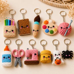May include: A collection of eleven small, crocheted keychains featuring various designs including a pencil holder, notebook, paintbrush, paint palette, yarn ball, glue bottle, scissors, sewing machine, tape measure, ghost, and a black spider.