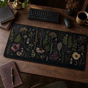May include: A rectangular desk mat featuring a dark black background decorated with a botanical pattern of pressed flowers, green leaves, ferns, and various crystals. Small golden crescent moons and stars are scattered throughout the design.