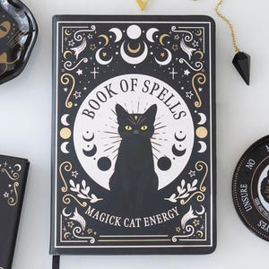 Peut inclure: Un carnet rectangulaire noir intitulé Book of Spells avec une illustration centrale d'un chat noir. La couverture présente des motifs célestes dorés et blancs, incluant des phases lunaires et des étoiles, avec le texte Magick Cat Energy.