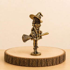 Puede incluir: Una figura de metal en tono dorado que representa a un personaje de pie con un sombrero de ala ancha y punta, un vestido con mangas abullonadas y botas. El personaje sostiene una escoba horizontalmente, con un pequeño libro rectangular.