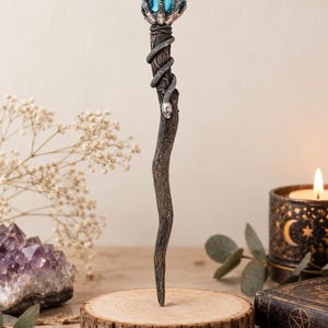 Op de afbeelding: Een decoratieve toverstaf met een donkere, getextureerde houten schacht omwikkeld door een zilverkleurige slang. De bovenkant toont een zilverkleurige klauw die een helderblauwe, gefacetteerde kristal vasthoudt. De schacht is gegraveerd met pentagrammen.