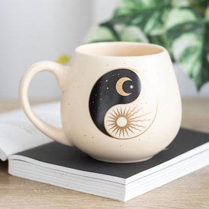 Könnte beinhalten: Ein cremefarbener Keramikbecher mit gesprenkelter Oberfläche und einem schwarz-goldenen Yin-Yang-Design auf der Vorderseite. Die Grafik zeigt einen goldenen Halbmond mit Sternen im schwarzen Bereich und eine goldene Sonne mit Strahlen im cremefarbenen Bereich.