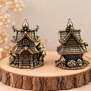 Op de afbeelding: Twee miniatuurhuisjes in messingkleur met gedetailleerde dakpannen, boogdeuren en kleine ramen. Elke structuur is aan de basis voorzien van decoratieve paddenstoelaccenten en heeft een puntige versiering op de top van het dak.