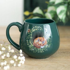 Peut inclure: Une tasse en céramique vert forêt foncé avec un corps arrondi et une anse. L'avant présente une illustration détaillée d'un renard orange endormi, recroquevillé dans une couronne circulaire de feuilles vertes, de fougères et d'un croissant de lune blanc.