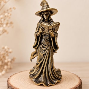Op de afbeelding: Een bronskleurig beeldje van een figuur met een puntmuts en een lange, zwierige jurk, die een open boek met ingewikkelde reliëfpatronen vasthoudt. Het beeldhouwwerk heeft een verweerde, metalen afwerking met donkere accenten in de plooien.