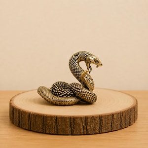 Peut inclure: Une petite figurine de serpent en métal doré, enroulée dans une posture défensive avec la tête relevée et la bouche ouverte. La surface du serpent présente des écailles texturées détaillées sur tout le corps, offrant un aspect réaliste.