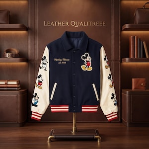Può includere: Una giacca varsity blu navy con maniche color crema, caratterizzata da una toppa di Topolino sul petto e molteplici grafiche di Topolino lungo le maniche. La giacca presenta polsini e orlo a coste a righe rosse e bianche, con la scritta "Mickey Mouse est. 1928" ricamata in oro sul davanti.