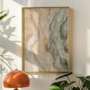 May include: A rectangular wall art piece in a gold frame featuring abstract fluid patterns in shades of beige, cream, and muted sage green with gold accents. The artwork is displayed above a table with a dome-shaped orange lamp and a green plant.