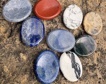 May include: A collection of nine smooth, polished oval stones in various colours and patterns, including solid red, deep blue with gold flecks, mottled grey, white with black veining, and dark green, arranged in a cluster on a textured surface.