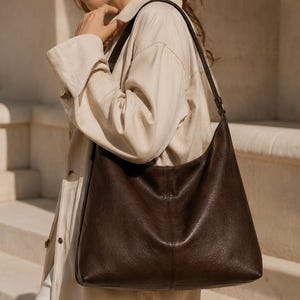 Puede incluir: Un bolso hobo de color marrón oscuro con una superficie texturizada suave y una sola correa para el hombro. El bolso presenta una costura vertical en el centro del panel frontal, una silueta holgada y un diseño minimalista sin herrajes visibles.