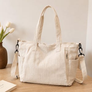 May include: A cream-colored corduroy tote bag featuring vertical ribbed texture, two top handles, and a detachable shoulder strap with black metal clips. The bag includes exterior side pockets and a vertical zippered pocket on the front panel.