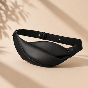 May include: A black crescent-shaped belt bag with a smooth, matte finish. The bag features a wide, integrated strap with a single visible gold-toned metal stud adjustment point and a structured, curved silhouette that tapers at both ends.