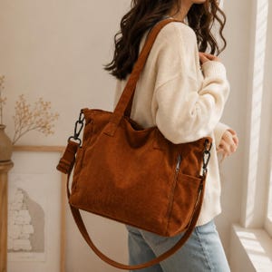 May include: A rust-coloured corduroy tote bag featuring a textured fabric surface, two top handles, and a detachable, adjustable shoulder strap with black metal hardware. The bag includes side pockets and a vertical zippered compartment on the side.