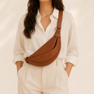 Pode incluir: Uma bolsa tiracolo em forma de meia-lua num tom castanho conhaque liso. A bolsa apresenta uma alça larga e ajustável com ferragens metálicas em tom dourado, usada a tiracolo sobre uma camisa branca e calças a combinar.