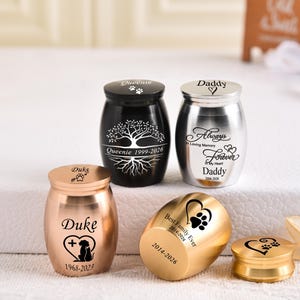 May include: A collection of five small metal cremation urns in black, silver, and gold finishes. Each urn features custom laser-engraved text and graphics, including names like Queenie, Daddy, and Duke, along with dates, paw prints, and heart motifs.
