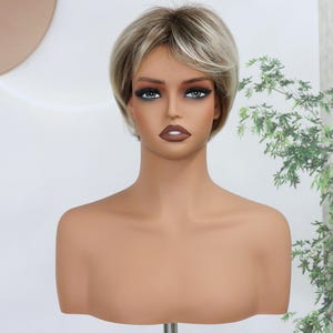 May include: A short, layered wig featuring a blend of ash blonde and light brown tones with darker roots. The hair is styled in a textured, voluminous pixie cut that frames the face, displayed on a smooth, beige-toned mannequin head and shoulders.