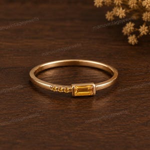 Può includere: Un sottile anello in oro caratterizzato da una singola pietra preziosa gialla rettangolare taglio baguette montata orizzontalmente, accentuata da tre piccole pietre gialle rotonde disposte in fila su un lato del gambo dell'anello.