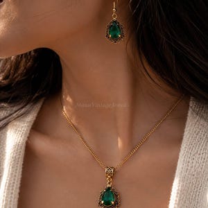 Puede incluir: Un conjunto de collar y pendientes colgantes en tono dorado con piedras en forma de l&aacute;grima de color verde esmeralda. El colgante y el pendiente est&aacute;n enmarcados por detalles de filigrana met&aacute;lica. El collar cuelga de una cadena dorada.