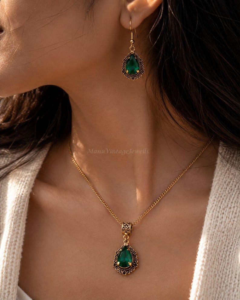 Puede incluir: Un conjunto de collar y pendientes colgantes en tono dorado con piedras en forma de l&aacute;grima de color verde esmeralda. El colgante y el pendiente est&aacute;n enmarcados por detalles de filigrana met&aacute;lica. El collar cuelga de una cadena dorada.