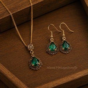 Puede incluir: Un conjunto de joyer&iacute;a en tono dorado que consta de un collar y un par de pendientes colgantes. Cada pieza presenta una piedra verde esmeralda en forma de l&aacute;grima engastada en un marco de metal ornamentado con filigrana. El collar incluye una cadena.
