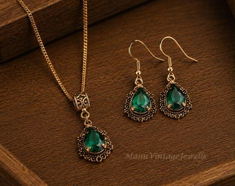 Puede incluir: Un conjunto de joyería en tono dorado que consta de un collar y un par de pendientes colgantes. Cada pieza presenta una piedra verde esmeralda en forma de lágrima engastada en un marco de metal ornamentado con filigrana. El collar incluye una cadena.