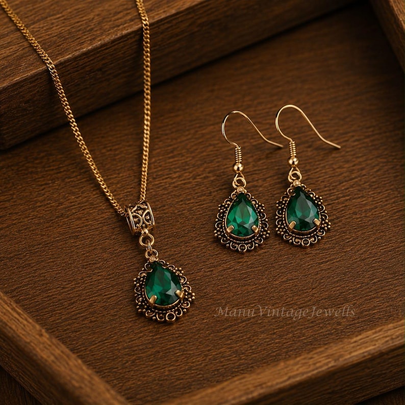 Puede incluir: Un conjunto de joyer&iacute;a en tono dorado que consta de un collar y un par de pendientes colgantes. Cada pieza presenta una piedra verde esmeralda en forma de l&aacute;grima engastada en un marco de metal ornamentado con filigrana. El collar incluye una cadena.