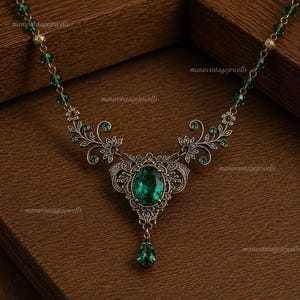 Puede incluir: Un collar con una piedra central ovalada de color verde esmeralda engastada en un marco de metal con tono antiguo y detalles de filigrana floral. Una pequeña piedra verde en forma de lágrima cuelga debajo, y la cadena incluye cuentas verdes y doradas.